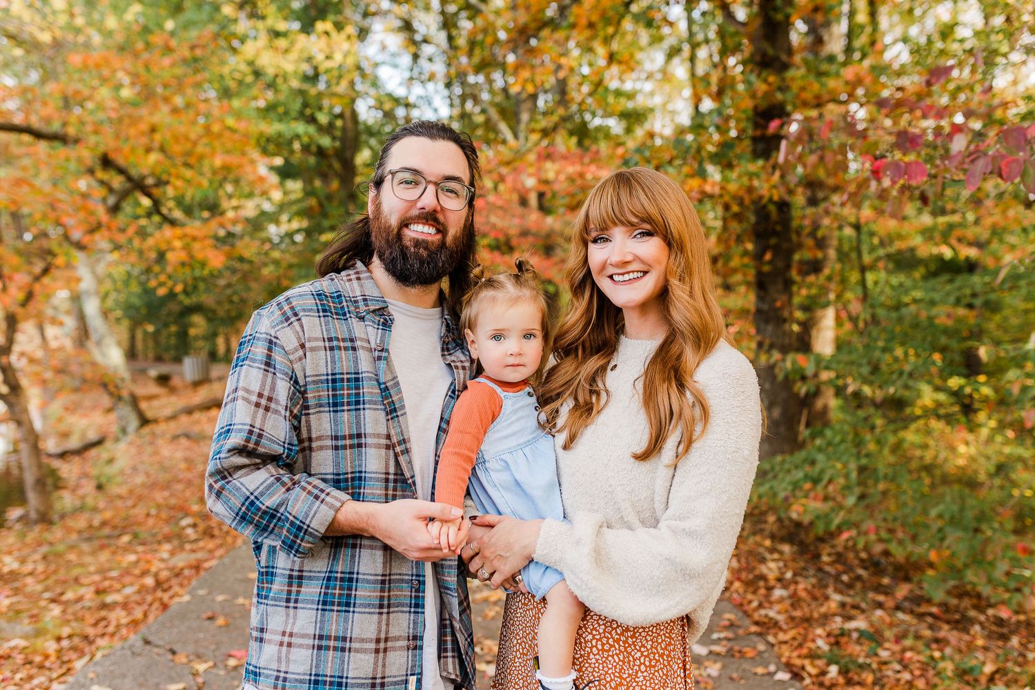 Three people smile together on a forest path surrounded by vibrant autumn foliage and fallen leaves.