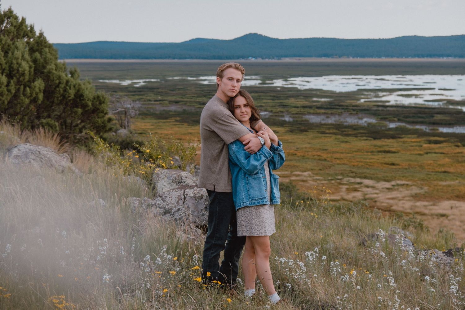 Two people embrace while standing on a grassy coastal hillside overlooking a scenic bay with mountains in the distance.