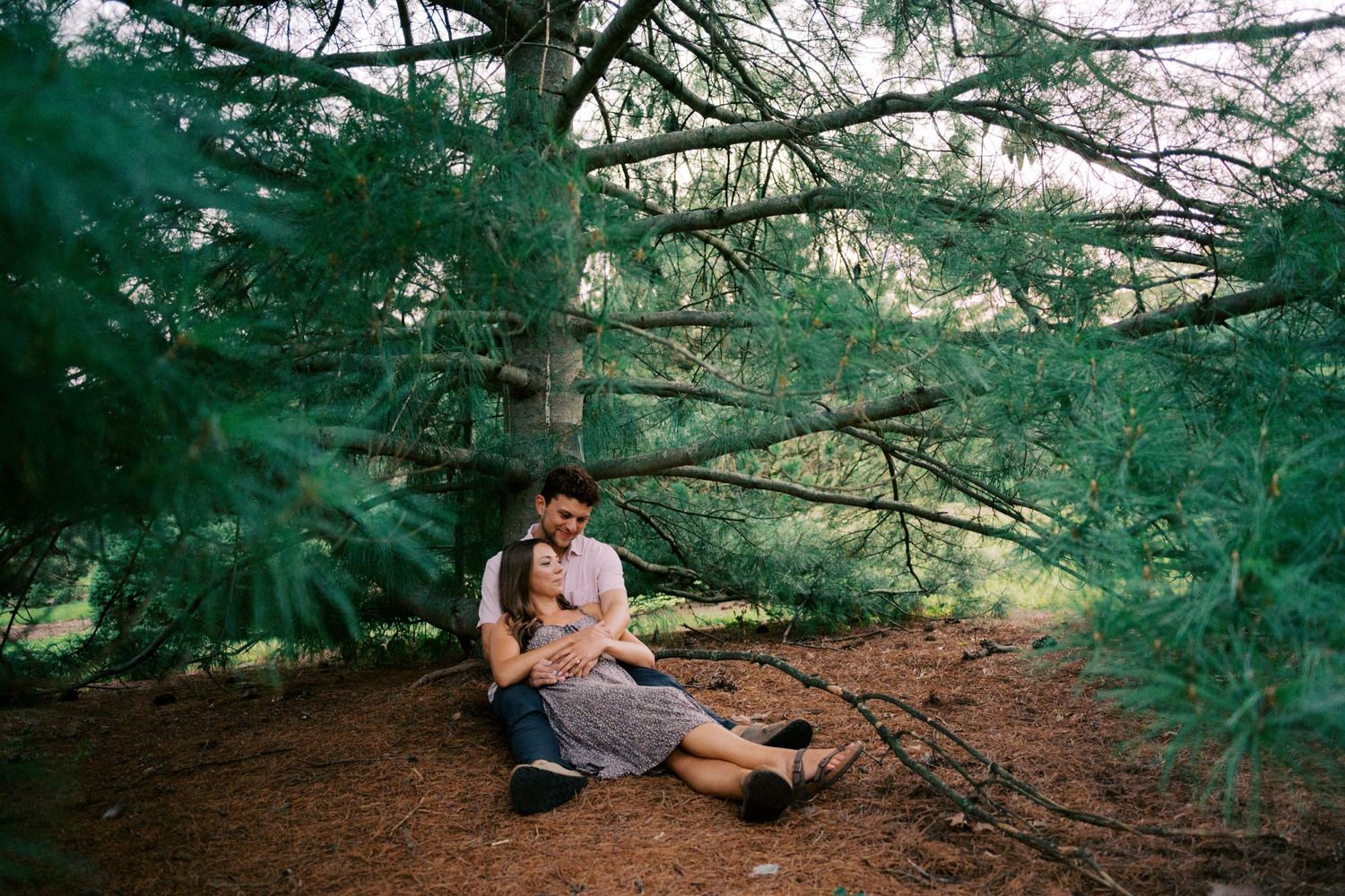 A couple sits on a forest trail beneath evergreen branches during an intimate moment in nature.