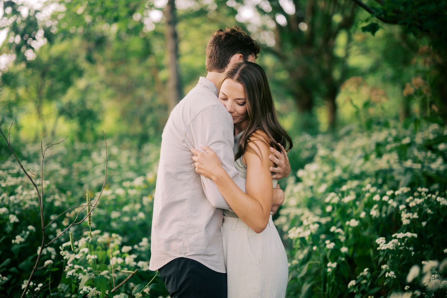 A romantic couple embraces tenderly among blooming white wildflowers in a lush garden setting.