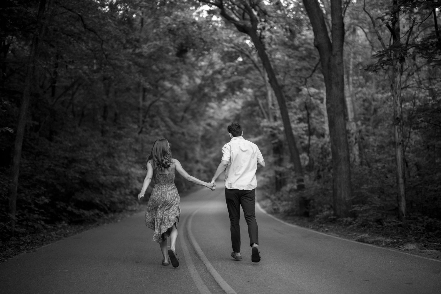 A couple walks hand in hand down a tree-lined road in black and white.