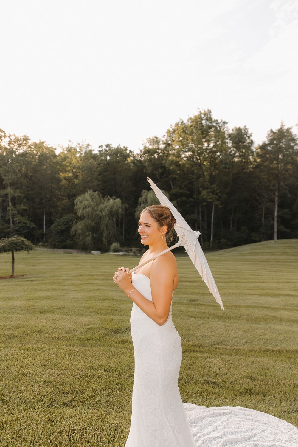 Bride in white lace gown holding elegant white parasol on lush green lawn with trees in background.