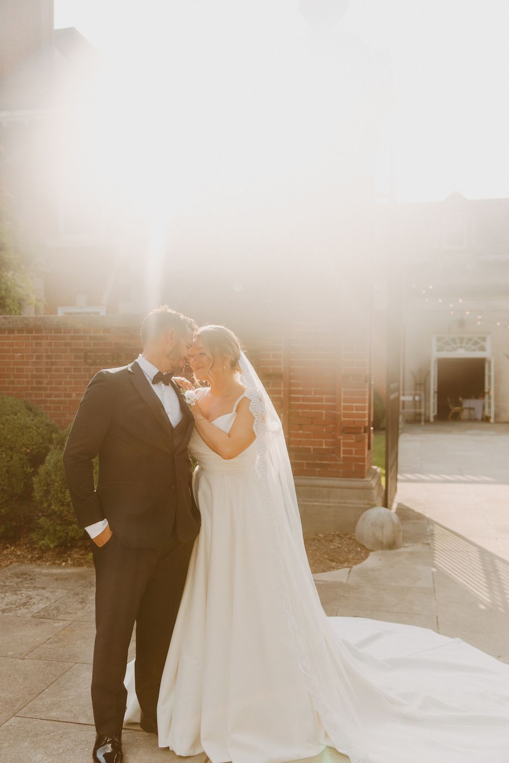 Bride and groom sharing a romantic moment in golden sunlight outside a brick building, bride in flowing white gown with veil.