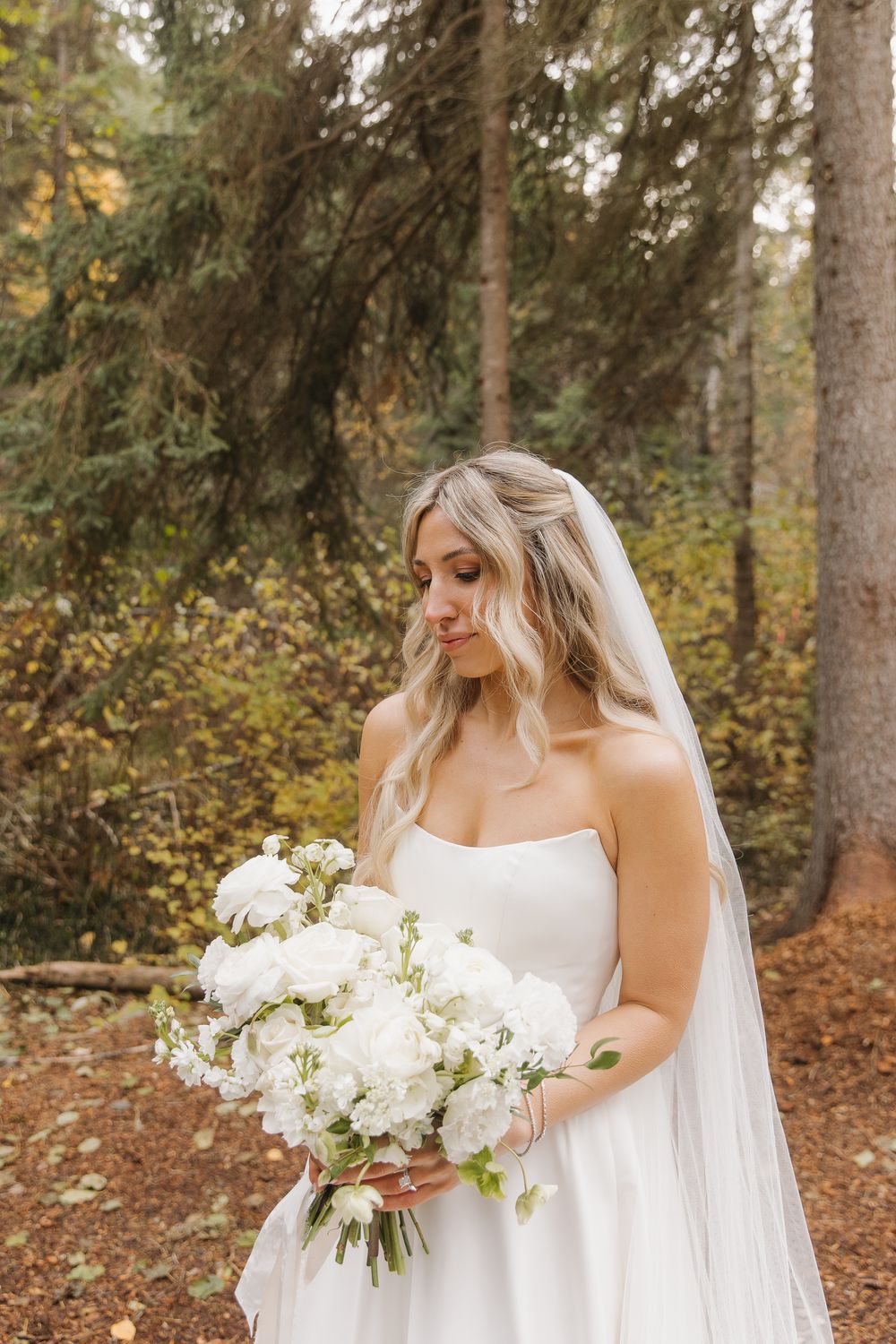Bride in flowing white wedding dress holds elegant white floral bouquet while standing in autumn forest setting in Kalispell, Montana.