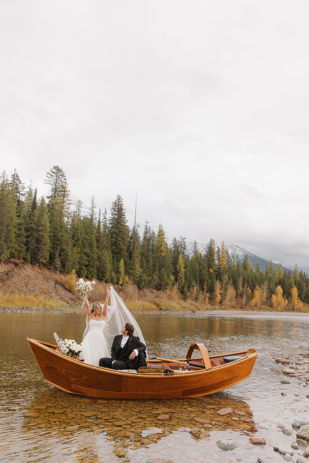 A couple sits together in a wooden drift boat on a scenic mountain lake surrounded by evergreen trees and autumn colors in Montana.