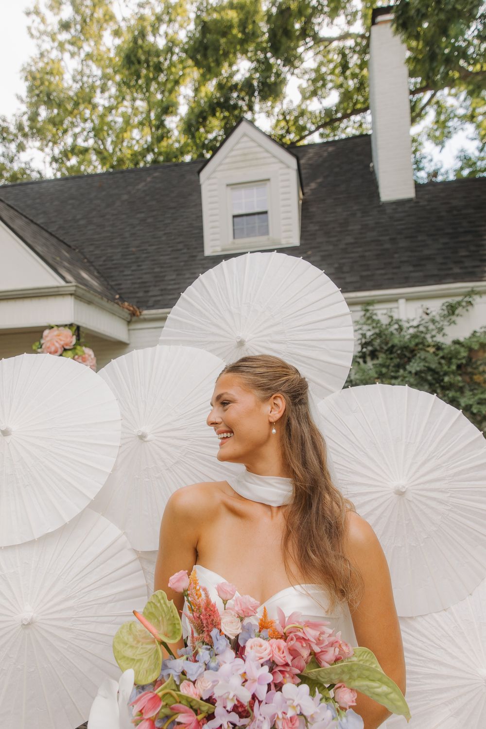 A bride in a white dress holds a pink bouquet while posing with decorative white paper parasols in an outdoor setting in Lexingotn, Kentucky