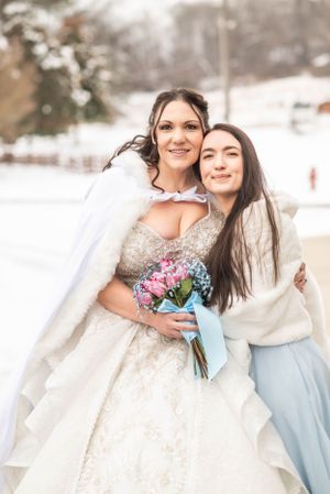 Winter bride in ballgown holds pink bouquet while embracing guest in snowy setting.