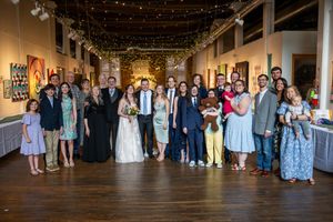 Wedding party and guests gather together in a rustic wooden-floored venue decorated with overhead string lights.