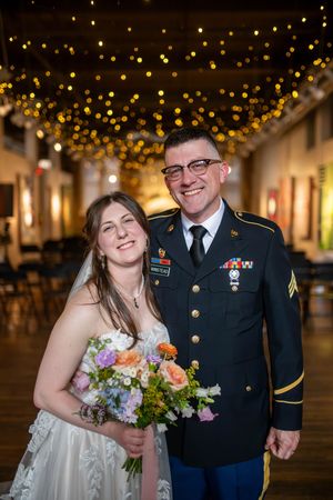 A bride and servicemember stand together under glowing string lights at their wedding reception.