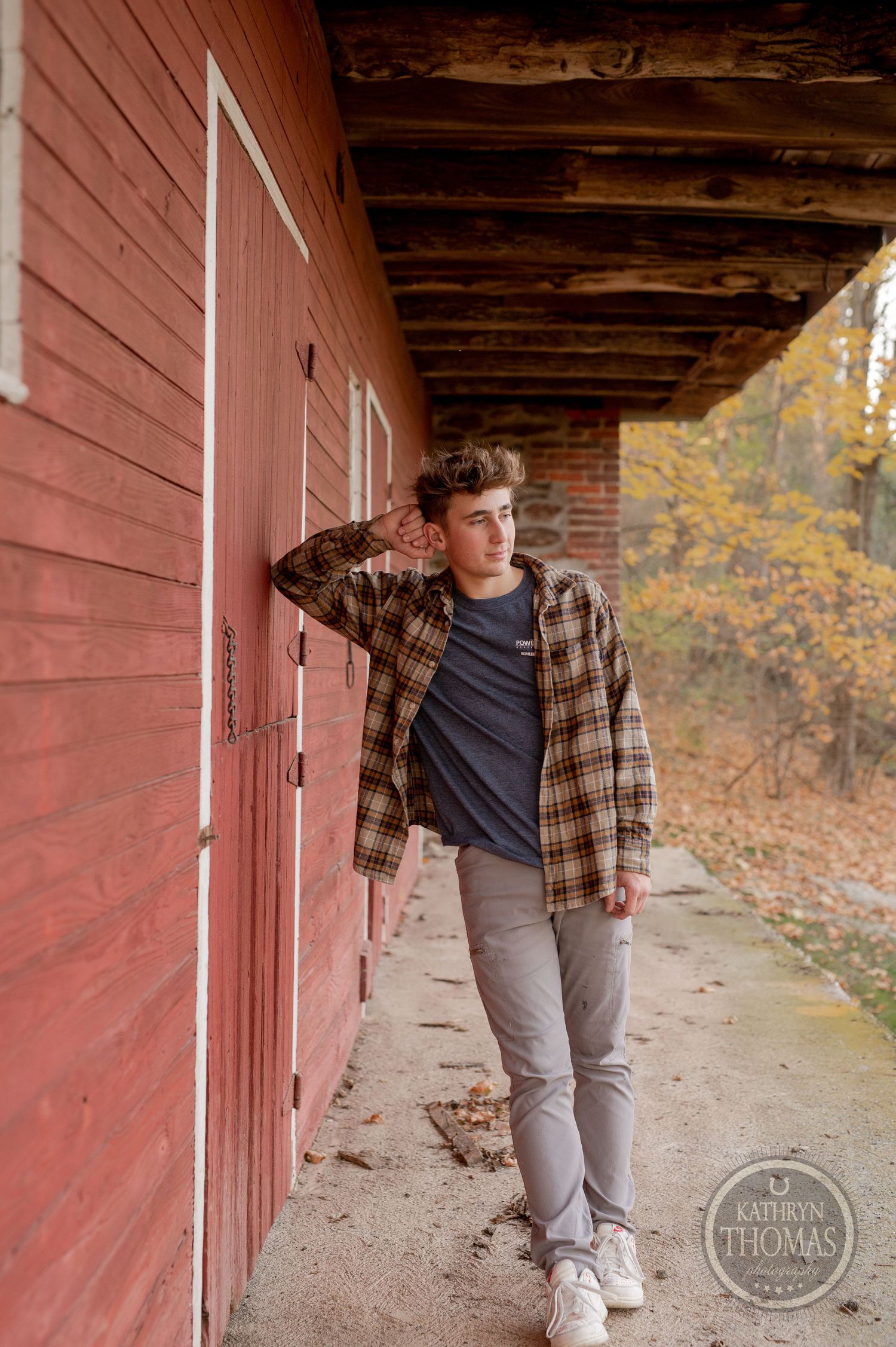 Someone stands in front of a red barn wearing a plaid shirt and gray pants on an autumn day.