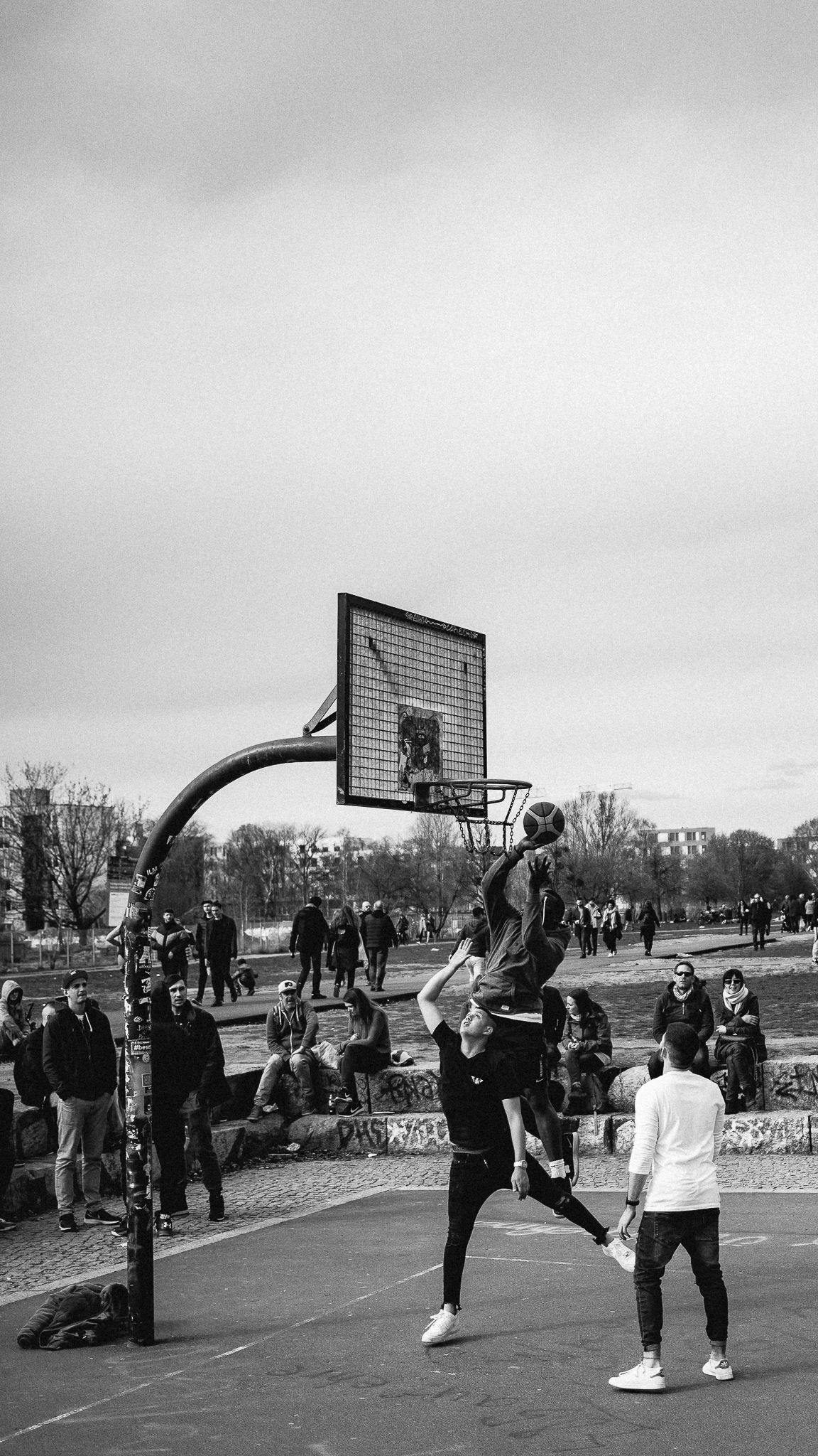 Basketball players competing at an outdoor court on a cloudy day while spectators observe the game.