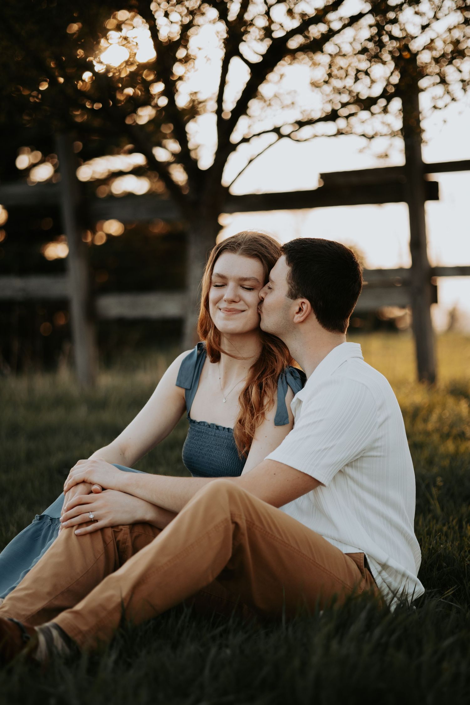 Couple sitting on grass at sunset, man kissing woman on cheek near rustic fence.