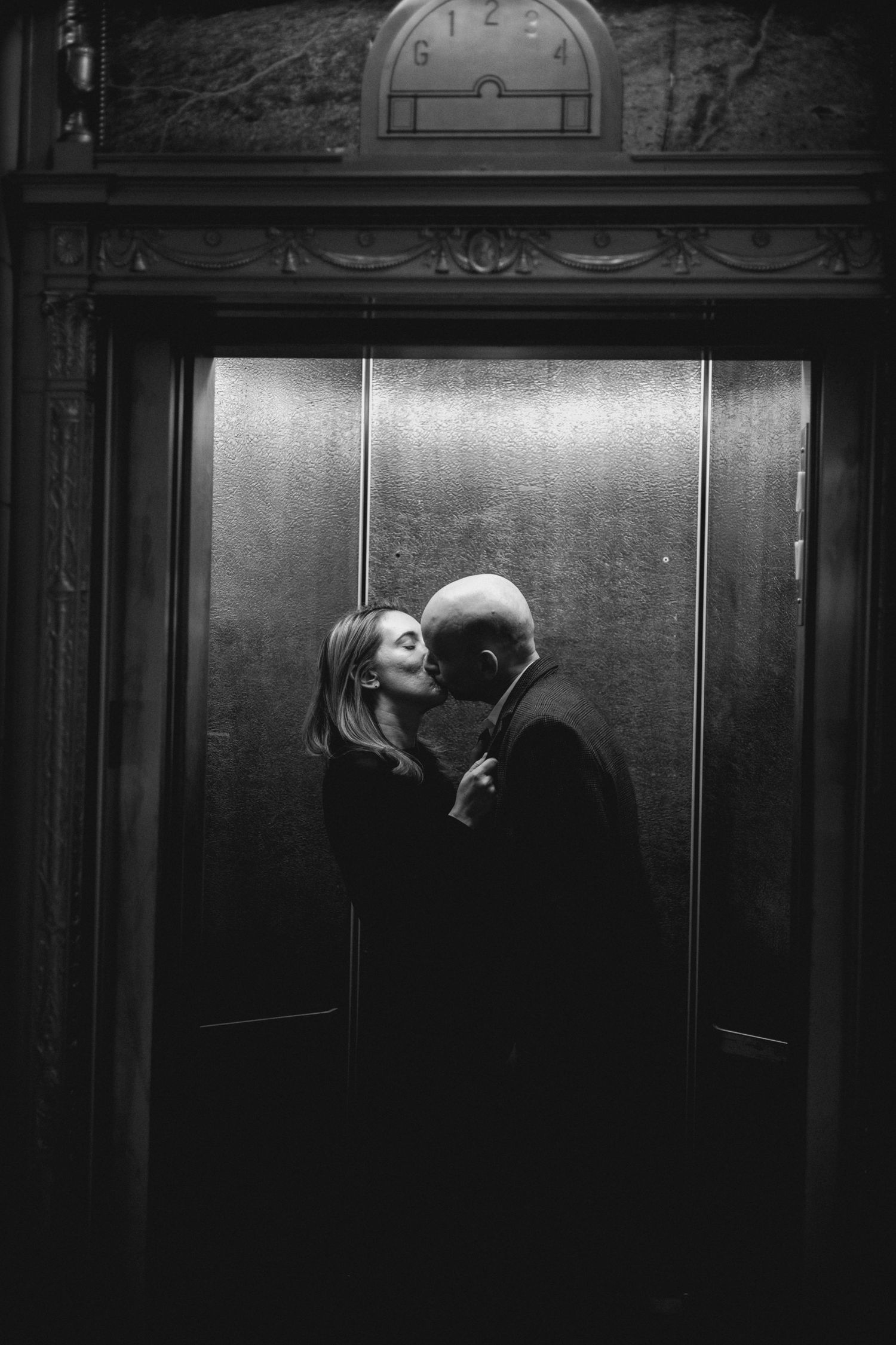 A black and white photograph shows two figures sharing an intimate moment in a dimly lit elevator. Hartford City Hall Engagement.