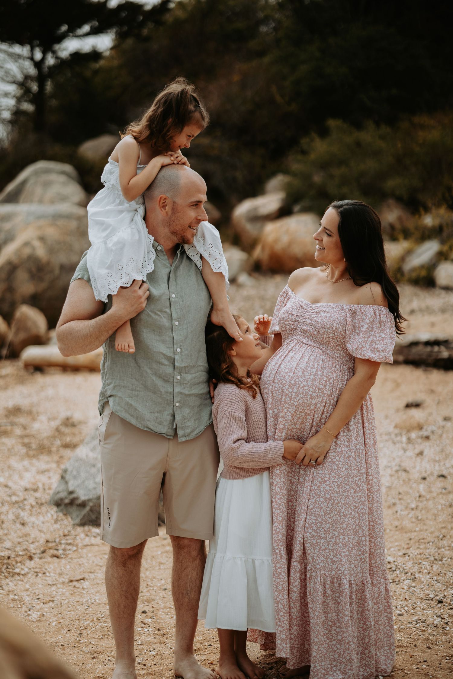 Pregnant woman in floral dress poses with husband and two young daughters on a rocky beach for a maternity photo.