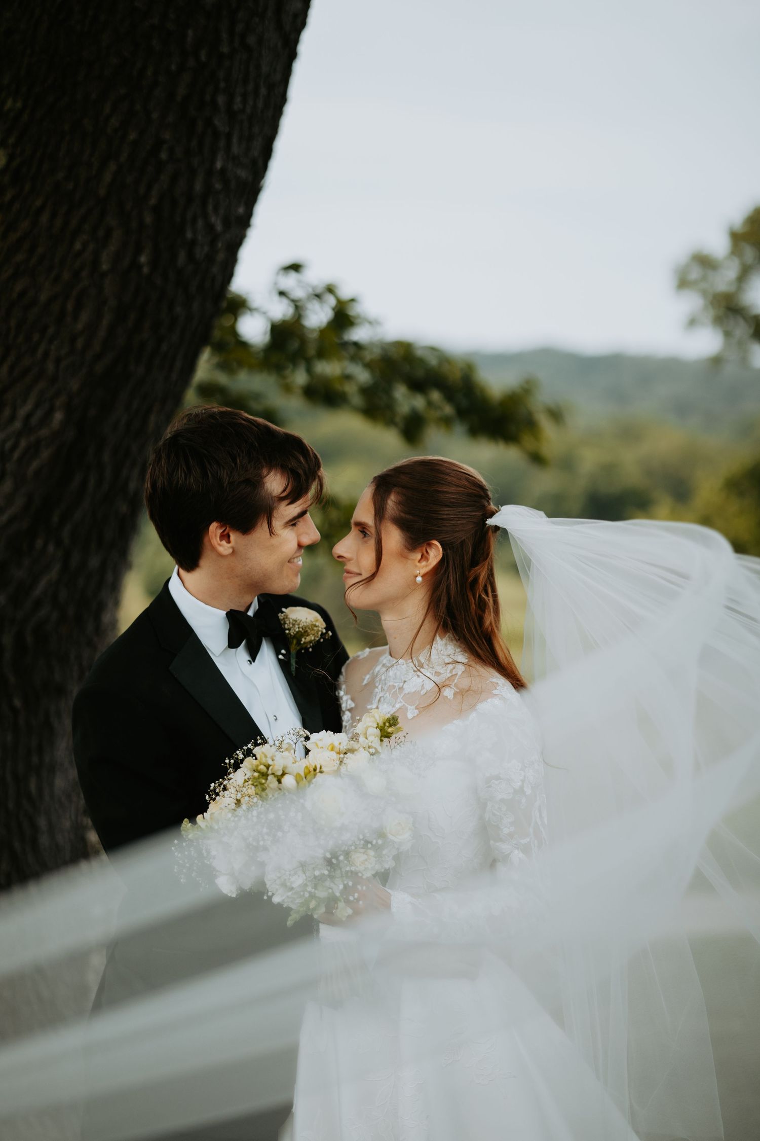 Bride and groom share a romantic moment outdoors, her veil flowing, as rolling green hills stretch behind them.