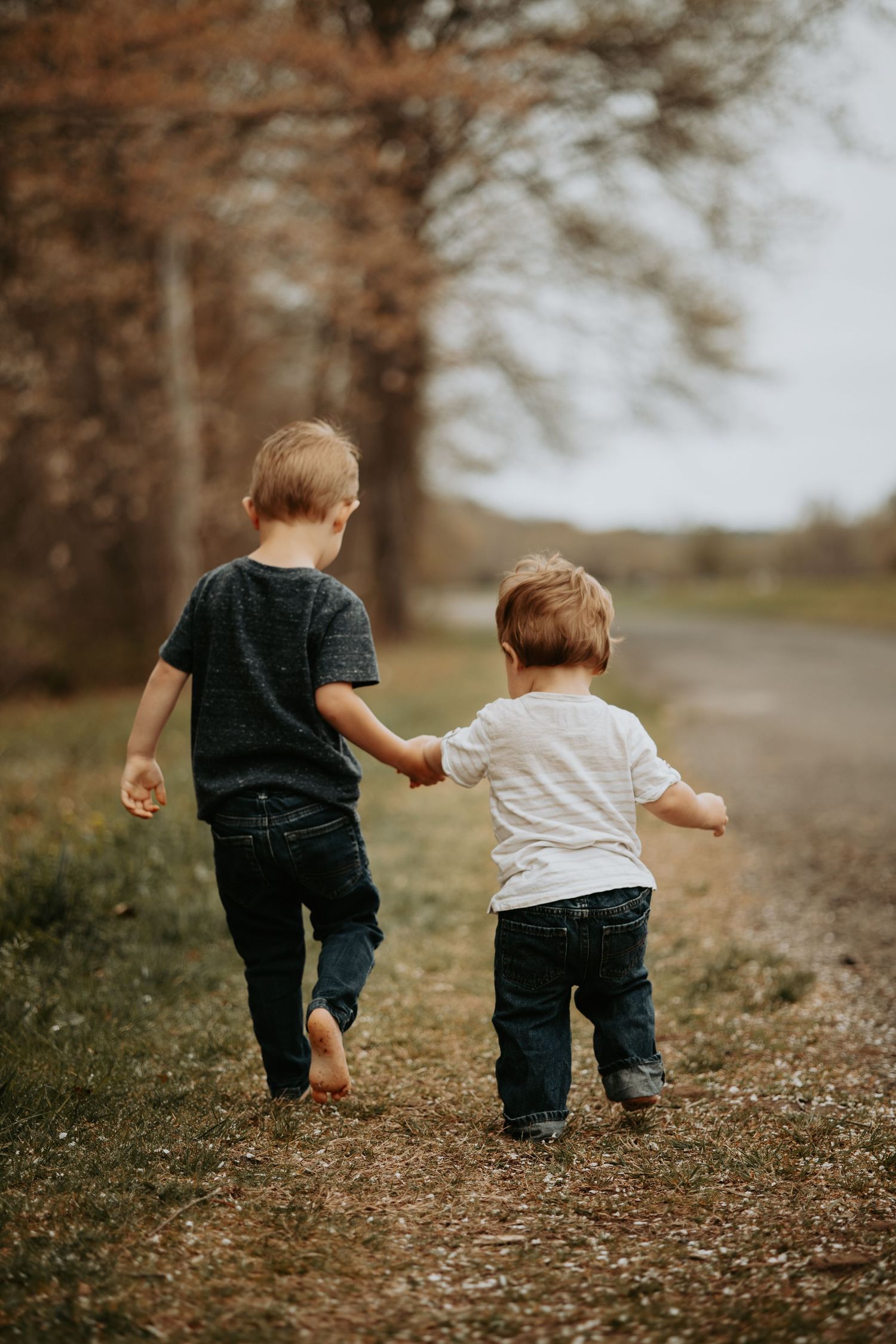 Two young boys holding hands while walking down a rural path surrounded by autumn trees.
