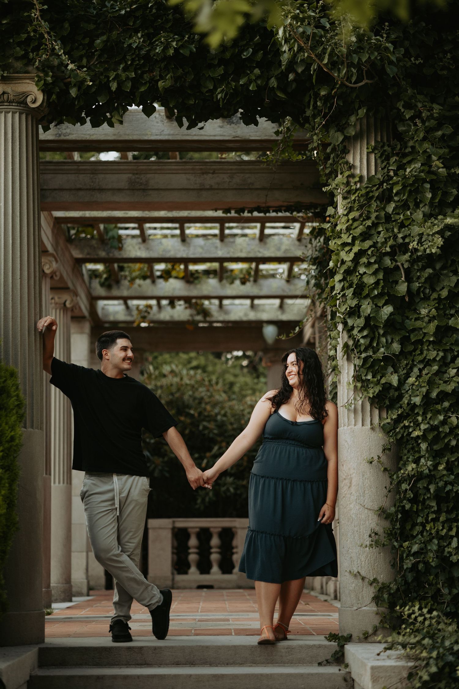 Couple holding hands and smiling under a vine-covered pergola with stone columns in a lush garden setting.