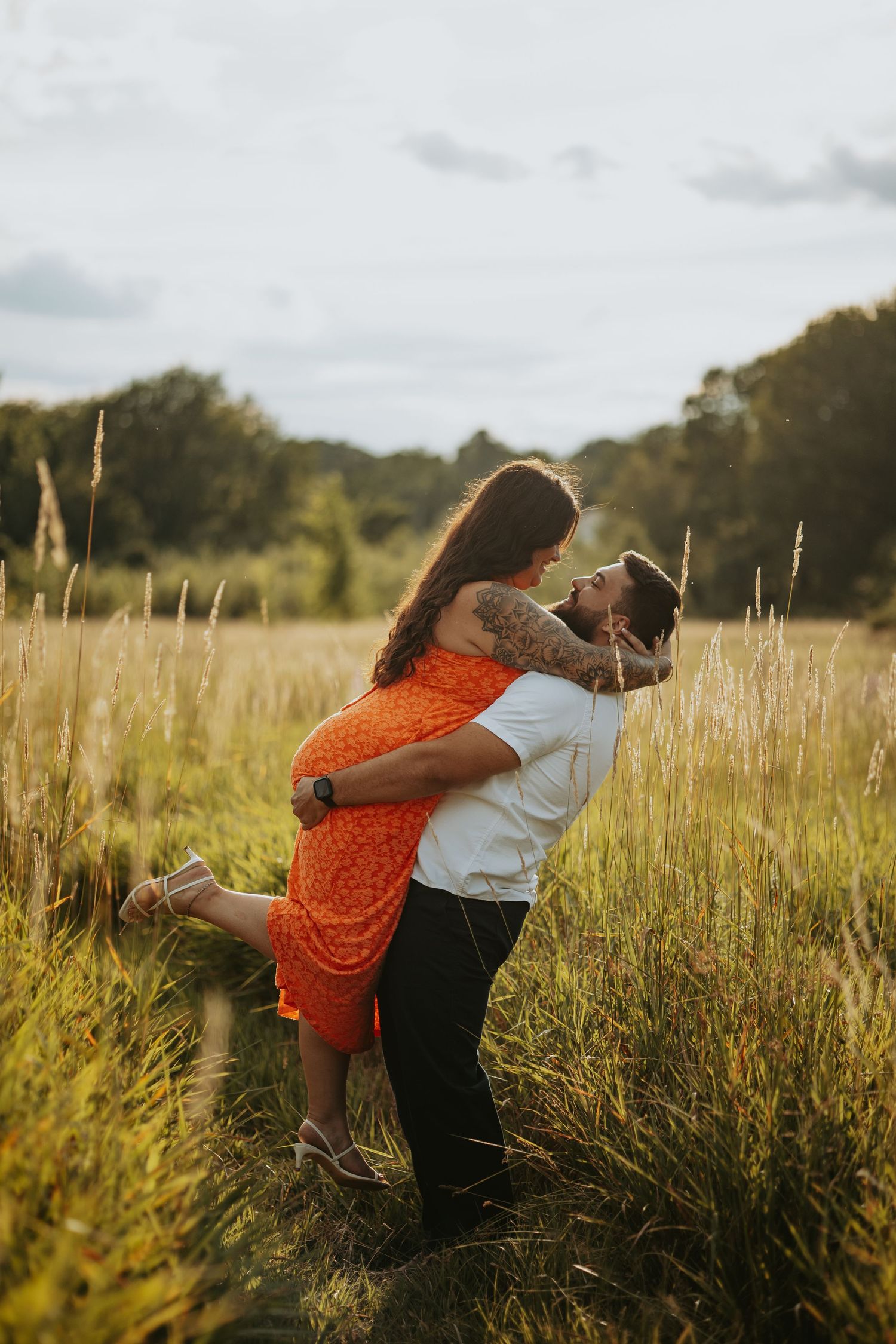 A couple shares a romantic embrace in a golden field, the man lifting the woman in an orange dress at sunset.
