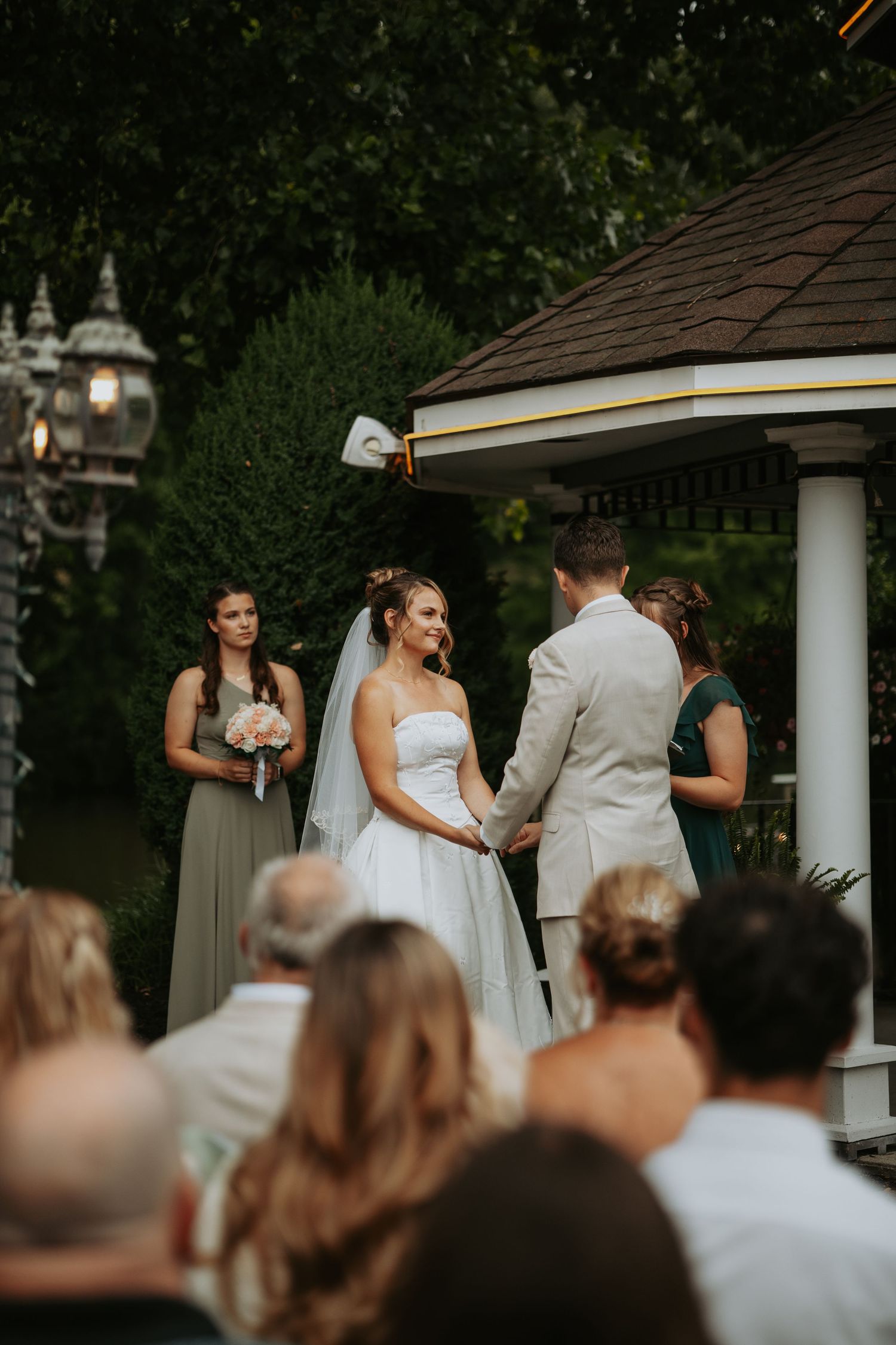 Bride and groom exchange vows at outdoor gazebo wedding ceremony with bridesmaids and guests watching.