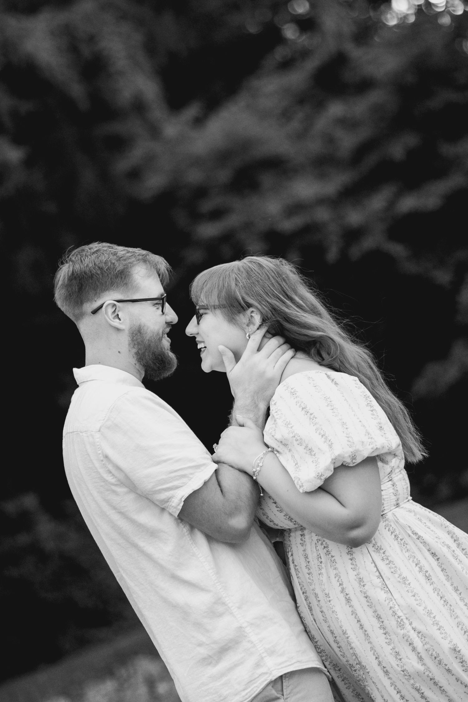 Black and white photo of a laughing couple embracing outdoors, man with glasses and beard, woman in dress, trees behind.