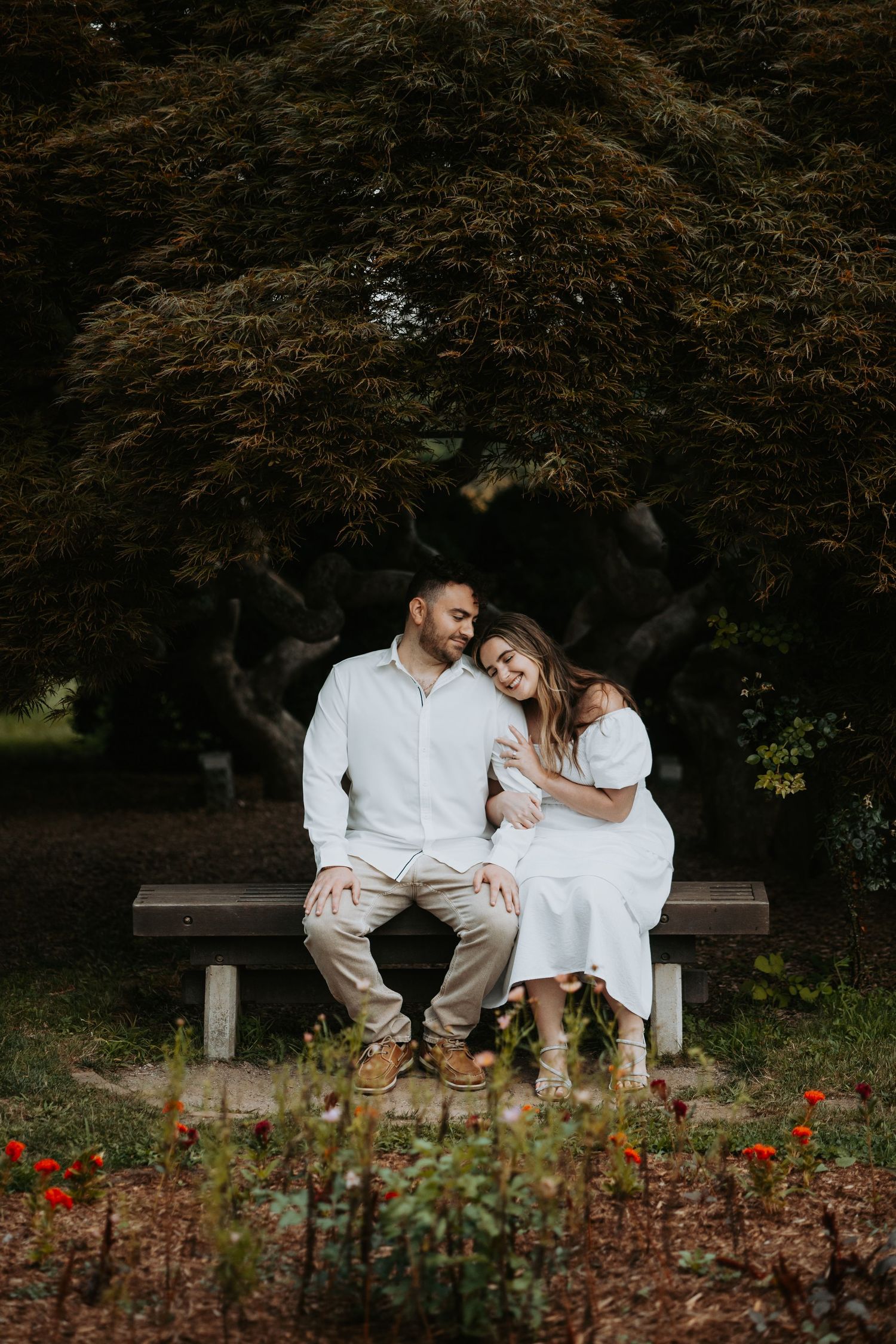Couple in white outfits sitting close together on a park bench surrounded by lush trees and red flowers.