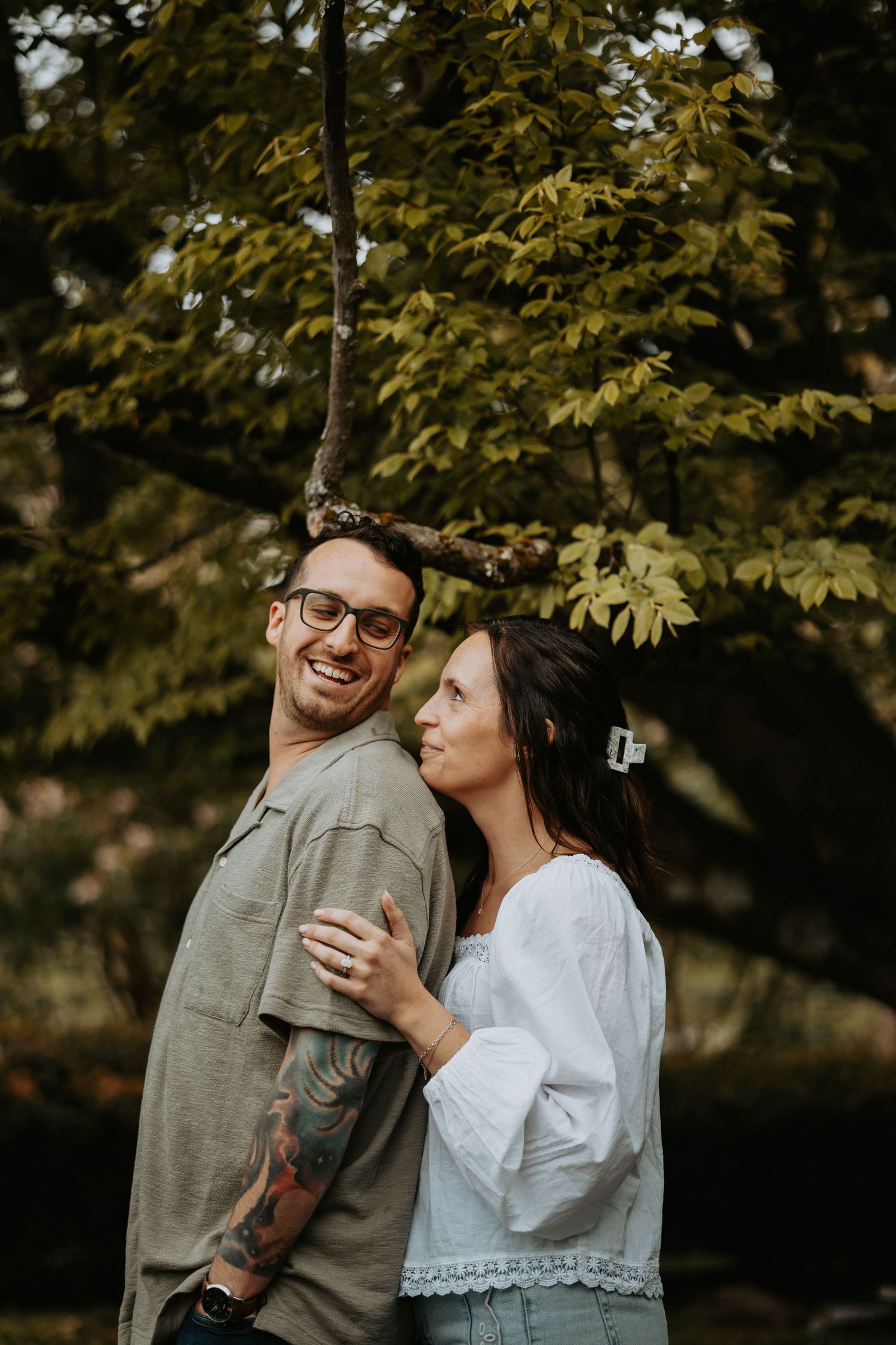 A smiling couple embraces outdoors under a lush green tree, man with tattoos and glasses, woman in white blouse.