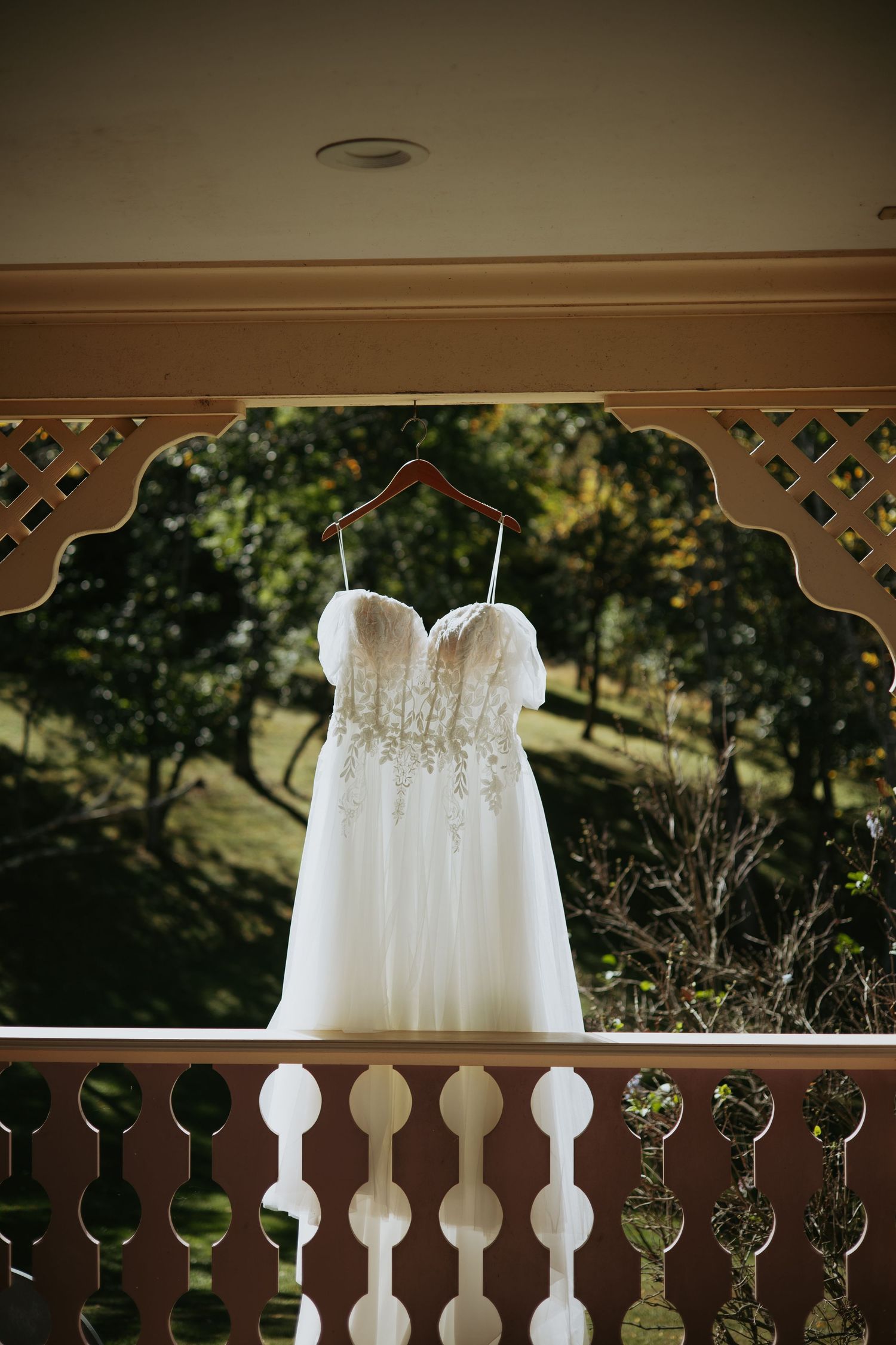 Strapless white lace wedding dress with beaded bodice hanging on a wooden hanger from a decorative porch overlooking sunlit trees.