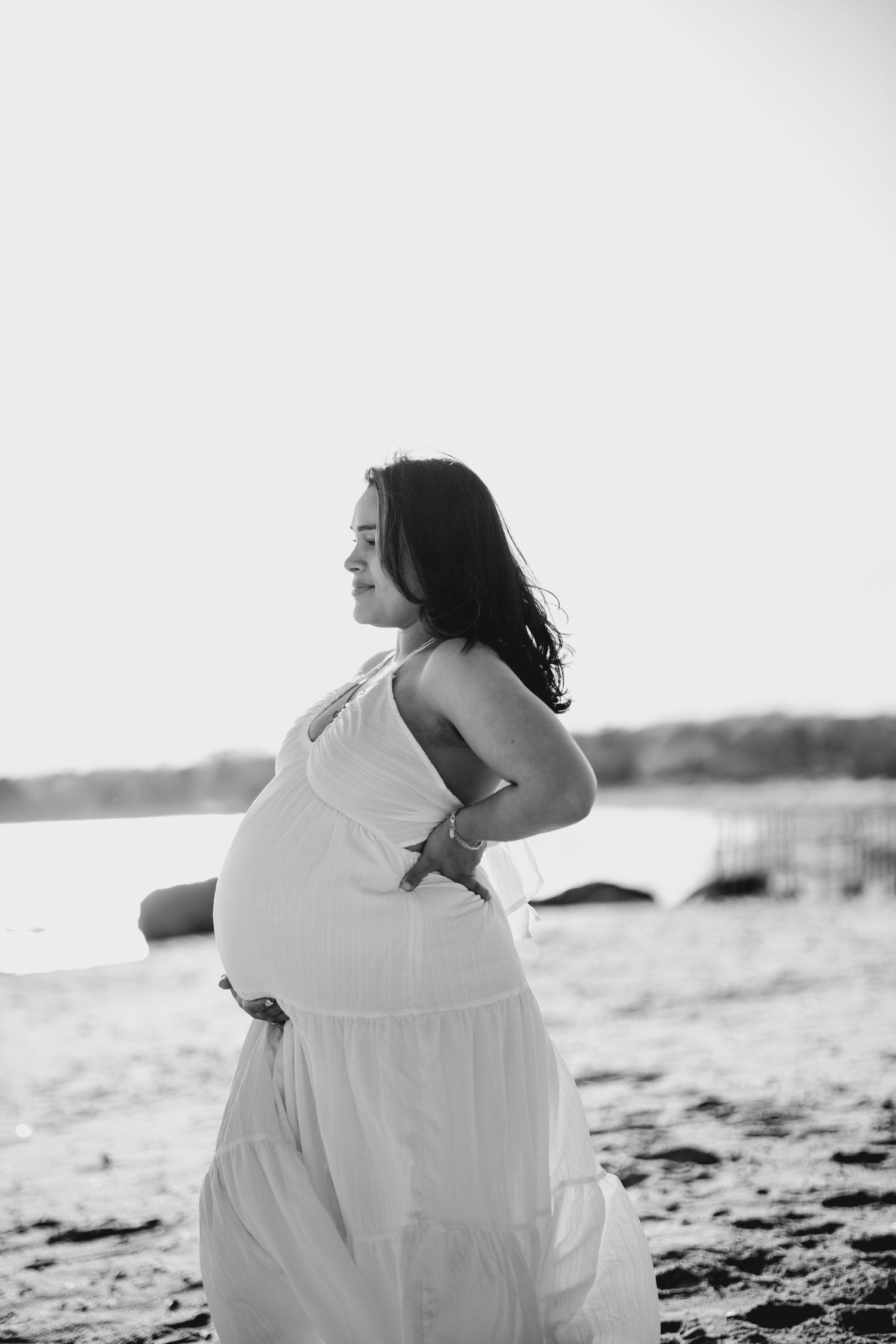 Black and white photo of a smiling pregnant woman in a white dress on a beach, cradling her baby bump.