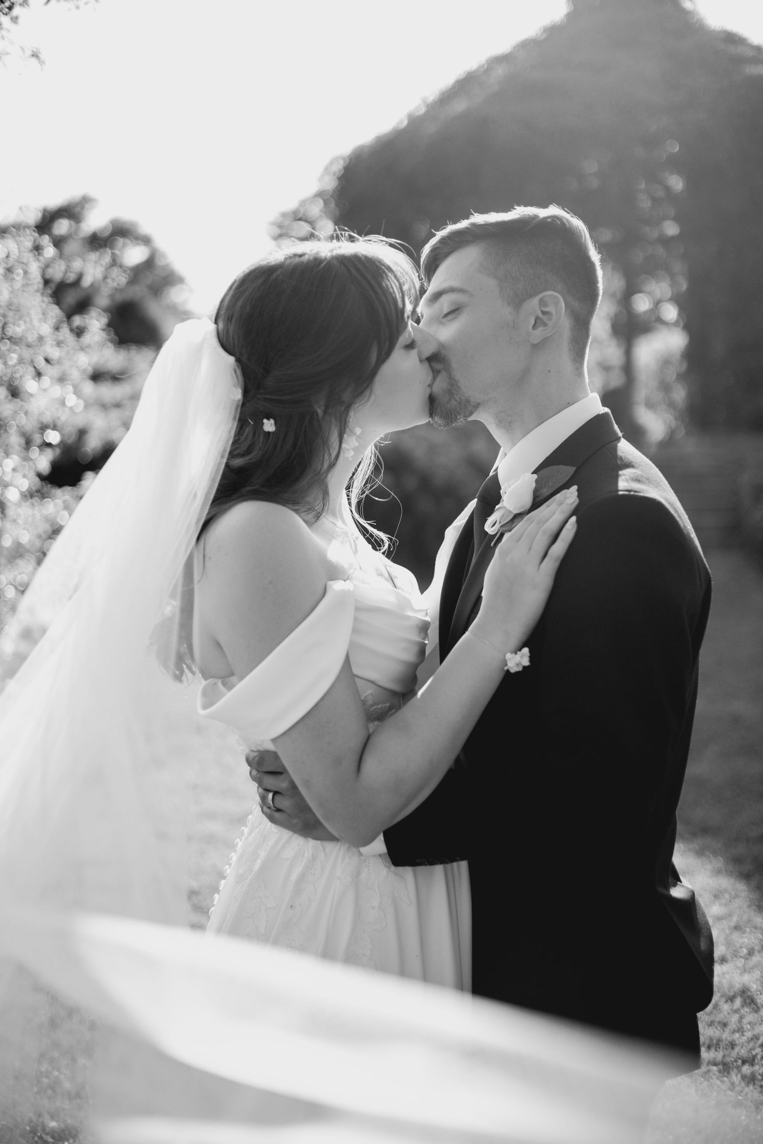 Black and white photo of a bride and groom sharing a romantic kiss outdoors on their wedding day.