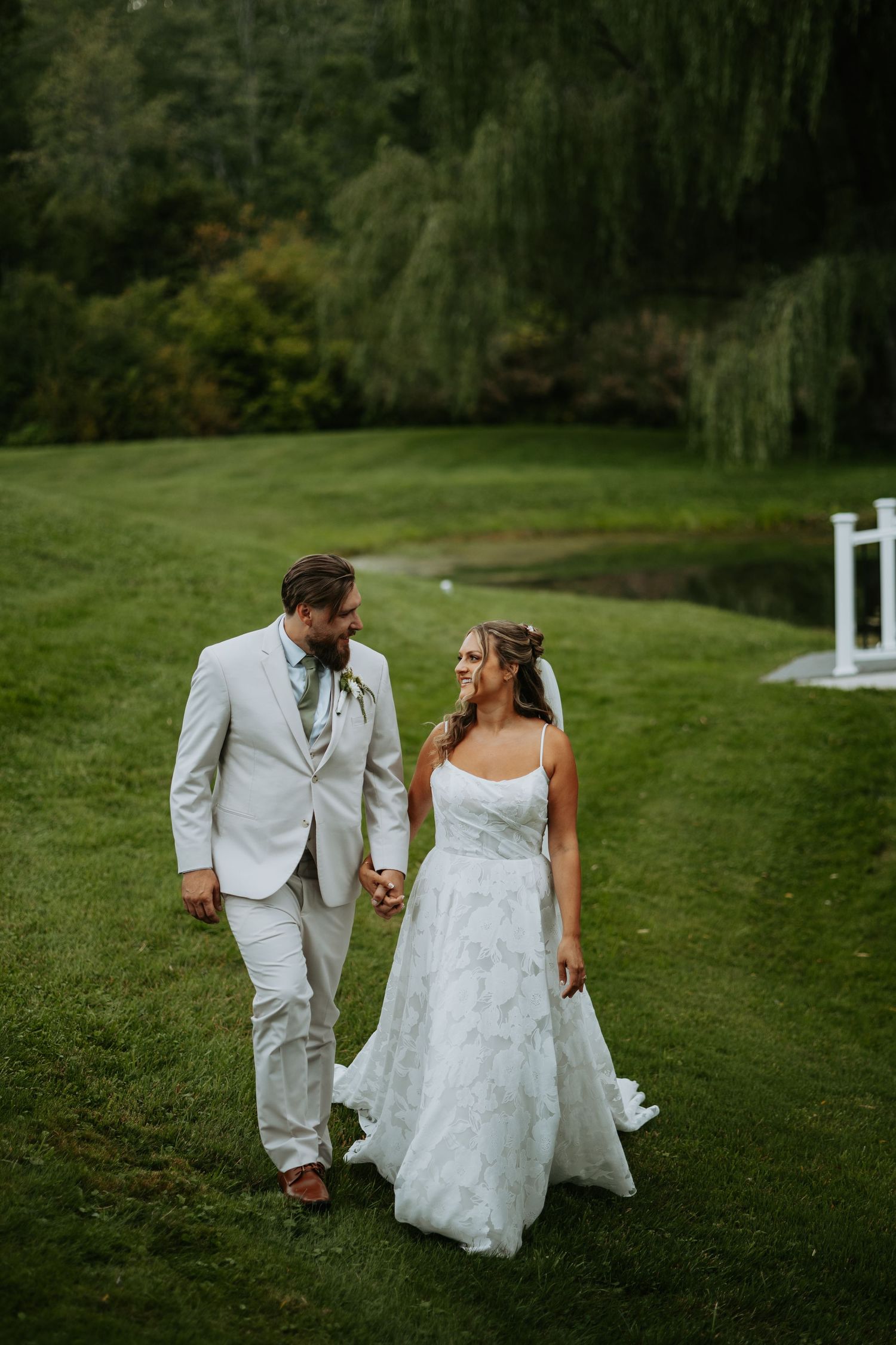 A bride and groom walk hand in hand across a lush green lawn, smiling at each other in elegant wedding attire.