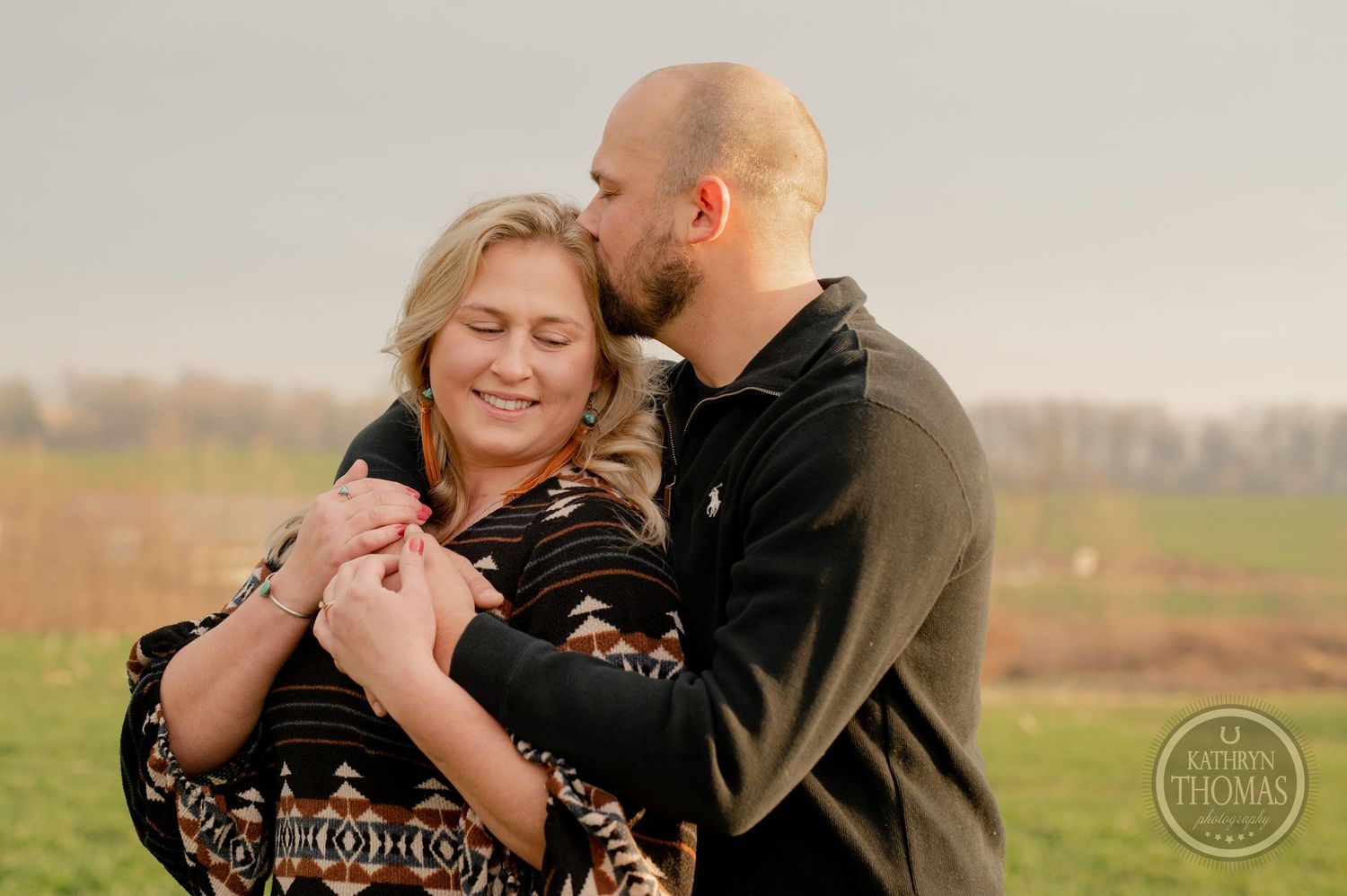 A romantic couple embraces in a field during golden hour wearing casual attire.
