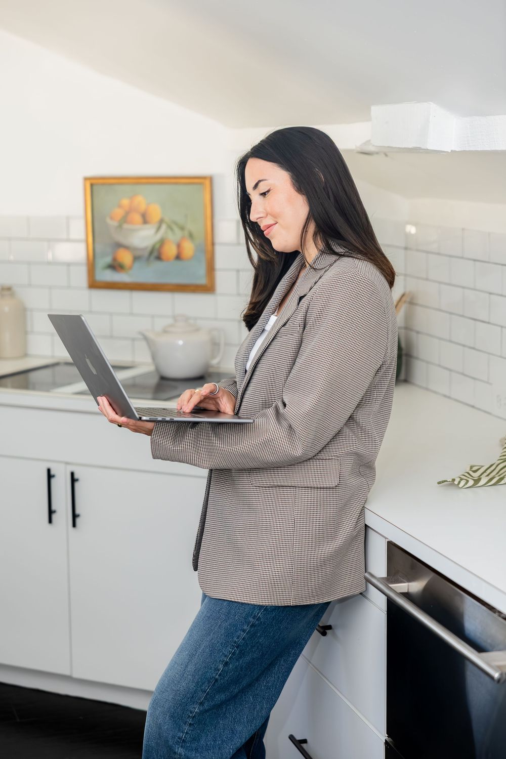 Woman in blazer and jeans using laptop in modern white kitchen with fruit painting on wall.