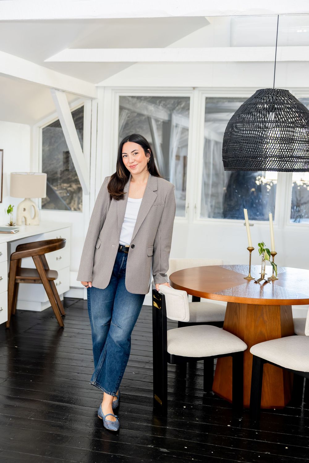 Woman in plaid blazer and jeans poses in modern dining room with dark wood floors and wicker pendant light.