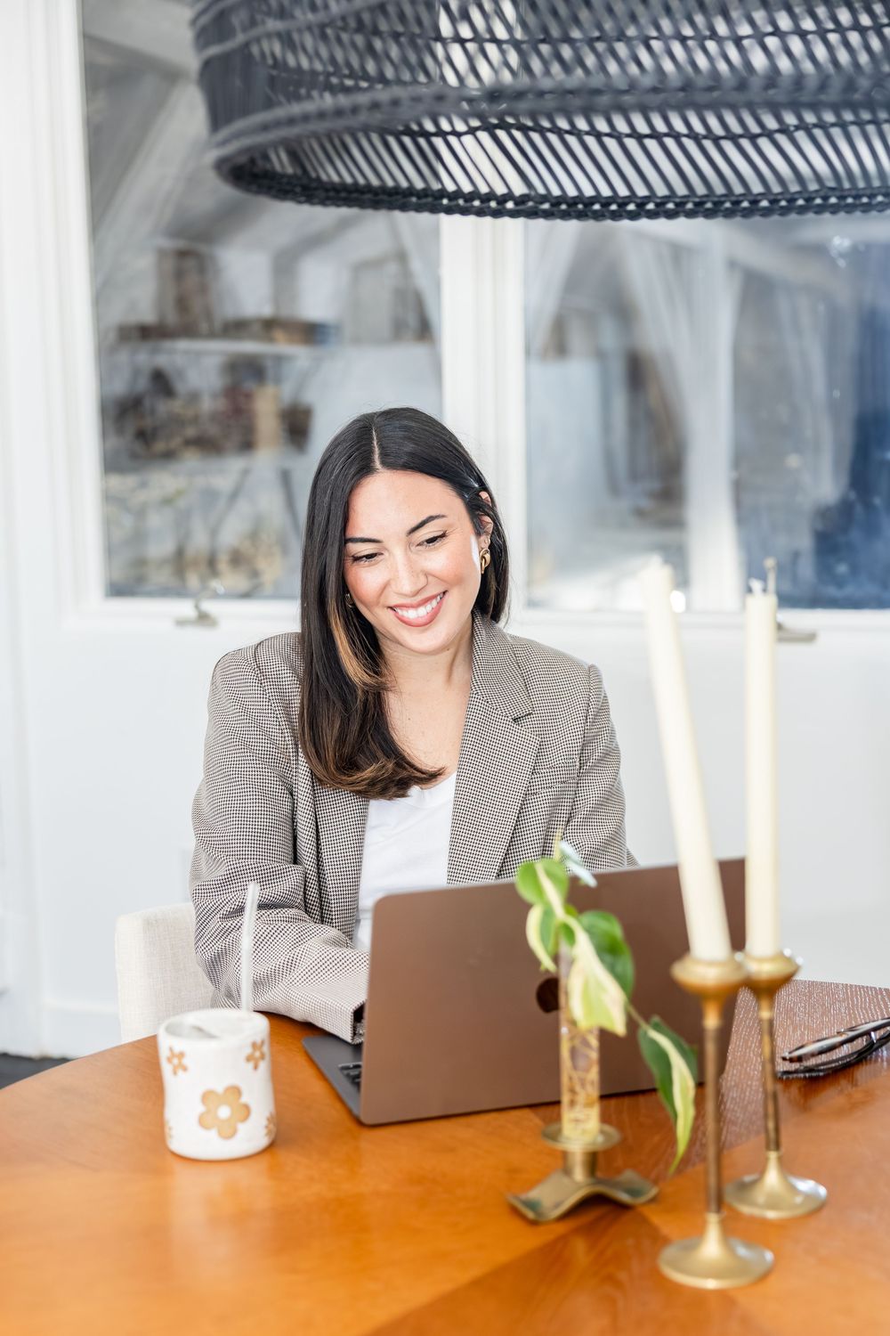 Smiling woman in blazer working on laptop at wooden desk with candles and coffee mug.