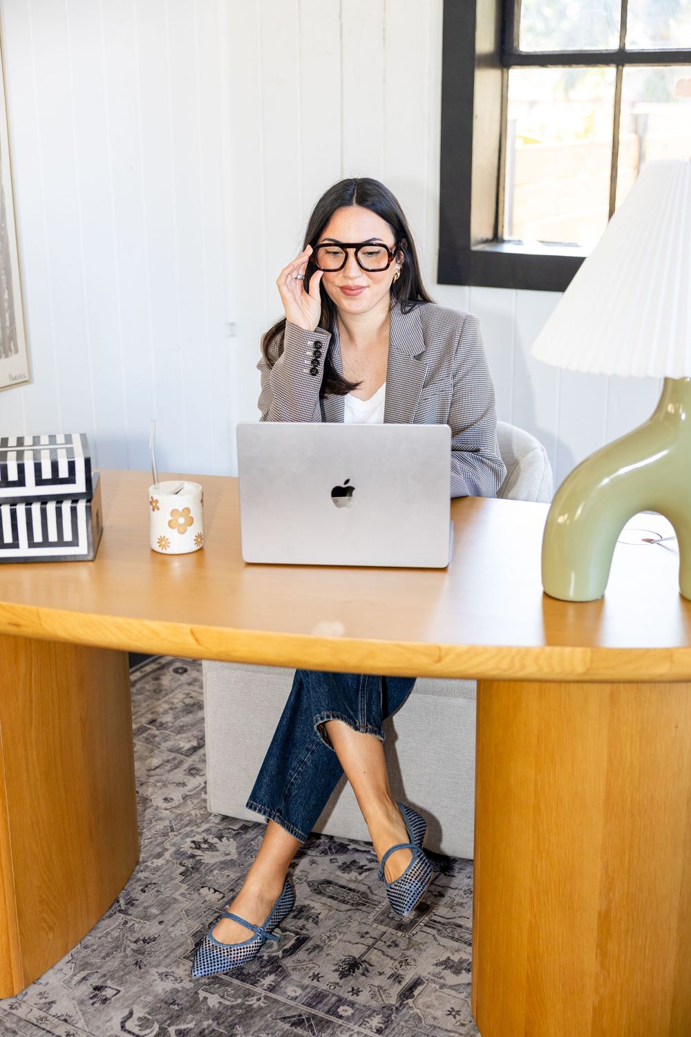 Woman in blazer and jeans working at wooden desk with MacBook laptop, adjusting glasses in stylish home office.