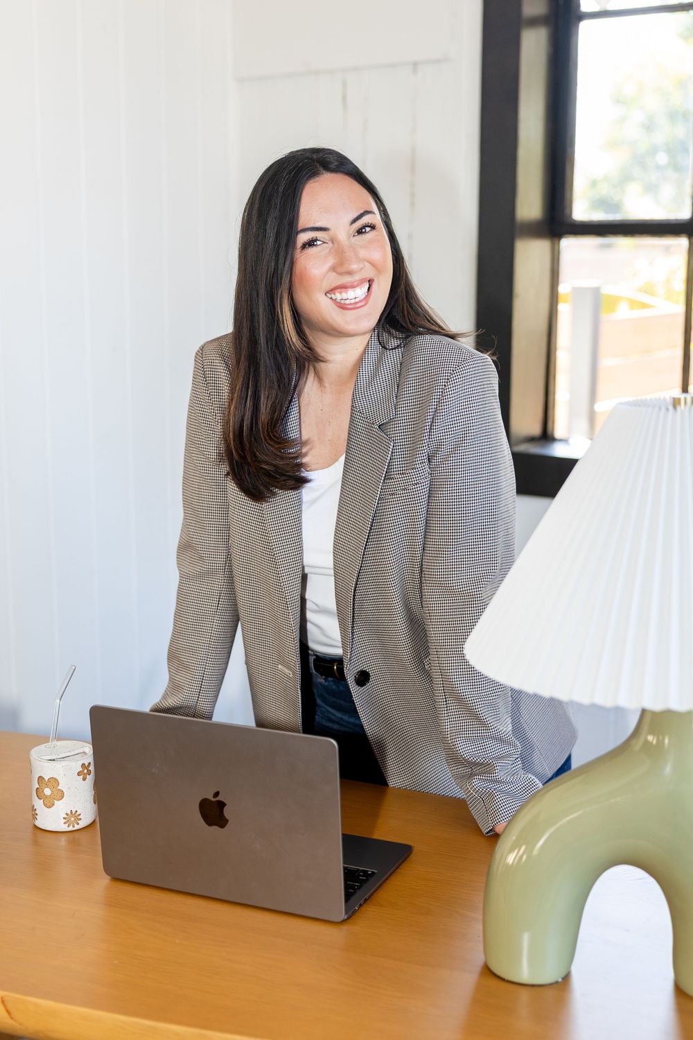 Smiling woman in gray blazer standing at wooden desk with MacBook laptop and green lamp in bright office.
