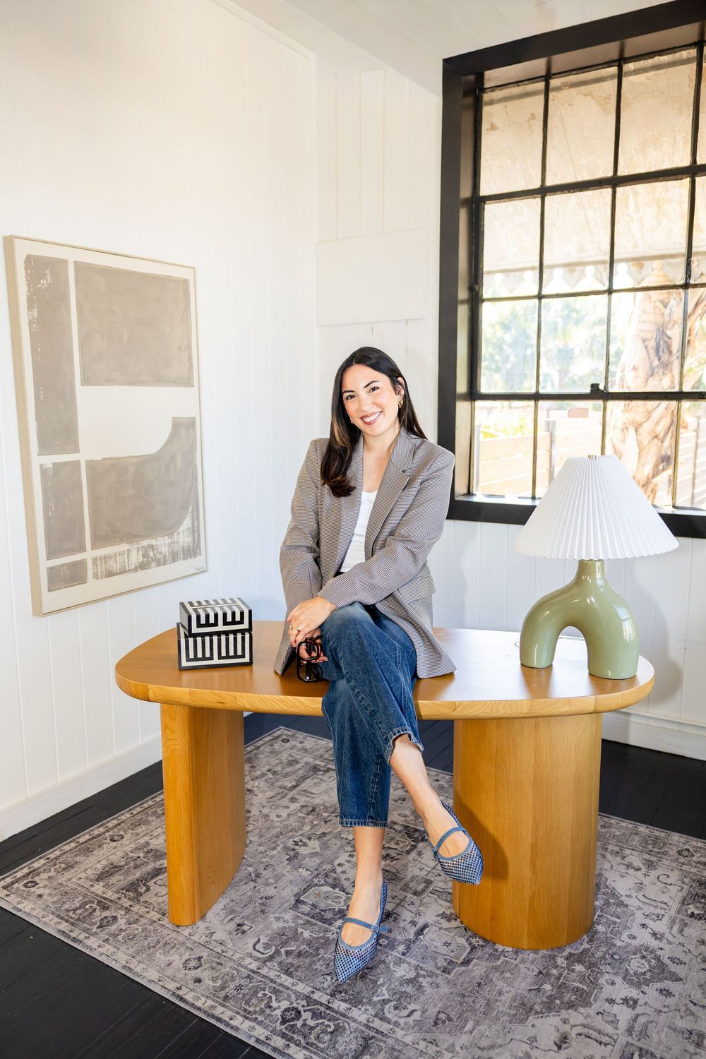 Stylish woman in blazer and jeans sitting on wooden round table near window.