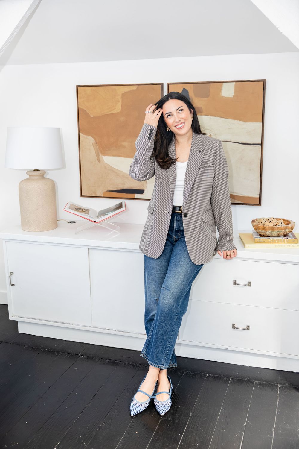 Woman in plaid blazer and jeans leaning against white cabinet with abstract art painting in bright home interior.