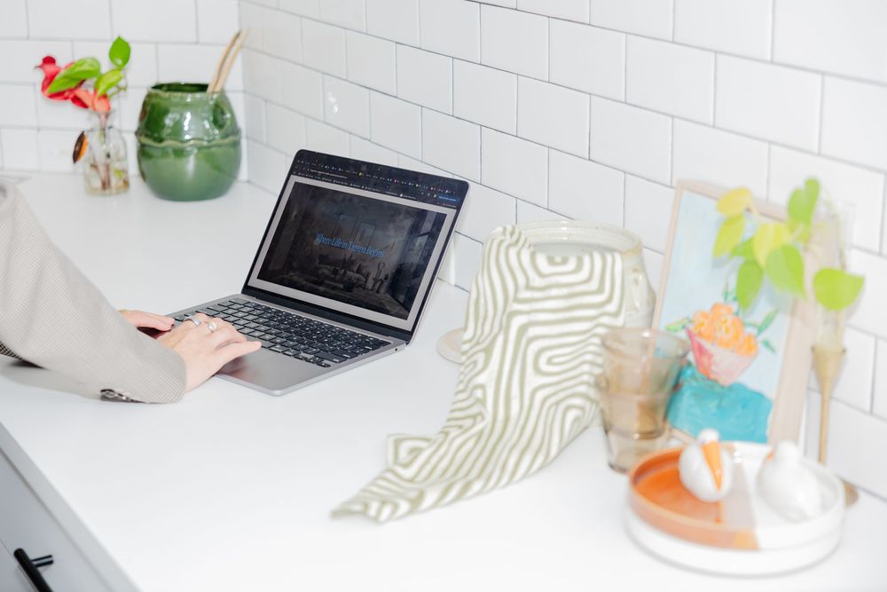 Person typing on laptop at a clean white desk with green vase, patterned cloth, and decorative items nearby.