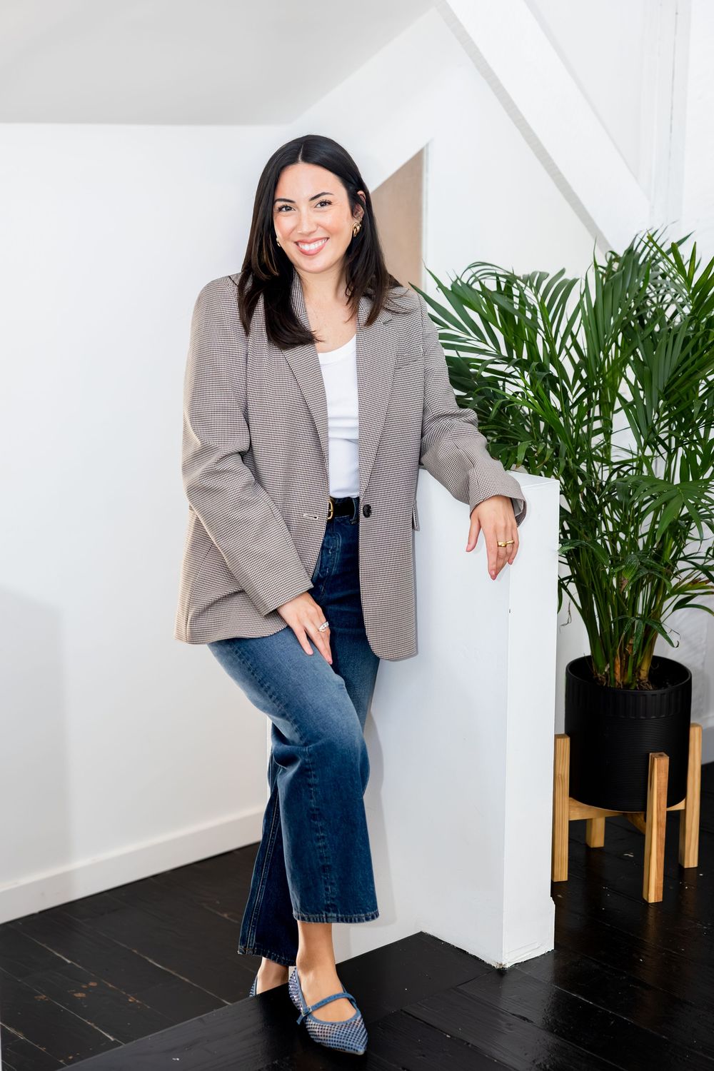Smiling woman in striped blazer and jeans leaning against white wall with tropical plant.