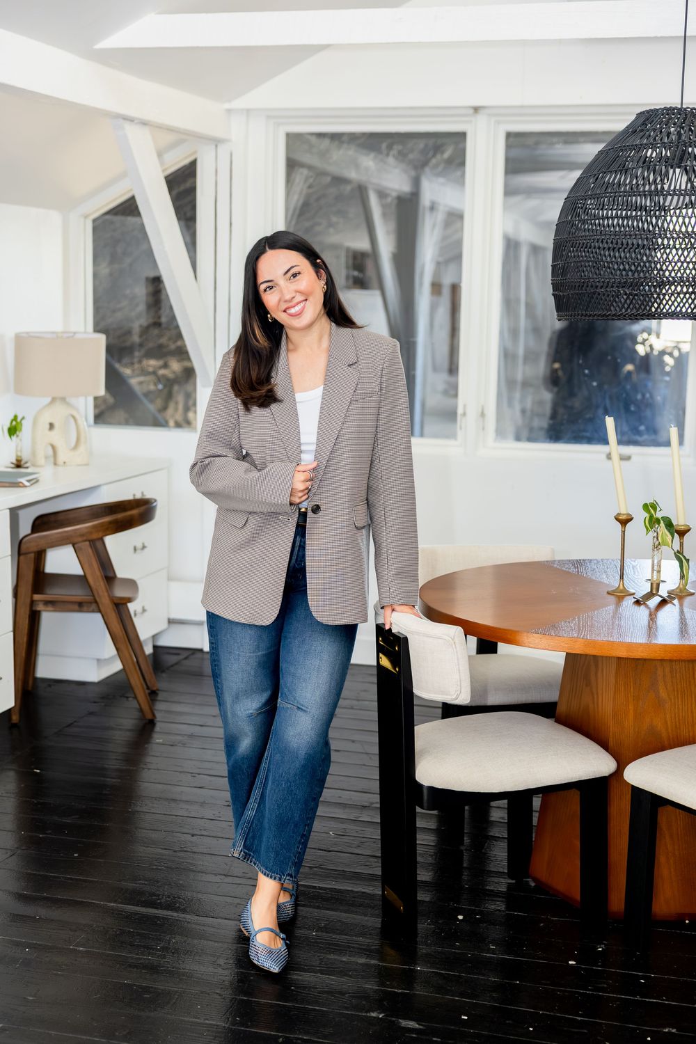 Smiling woman in gray blazer and jeans standing by round wooden dining table in bright modern home.