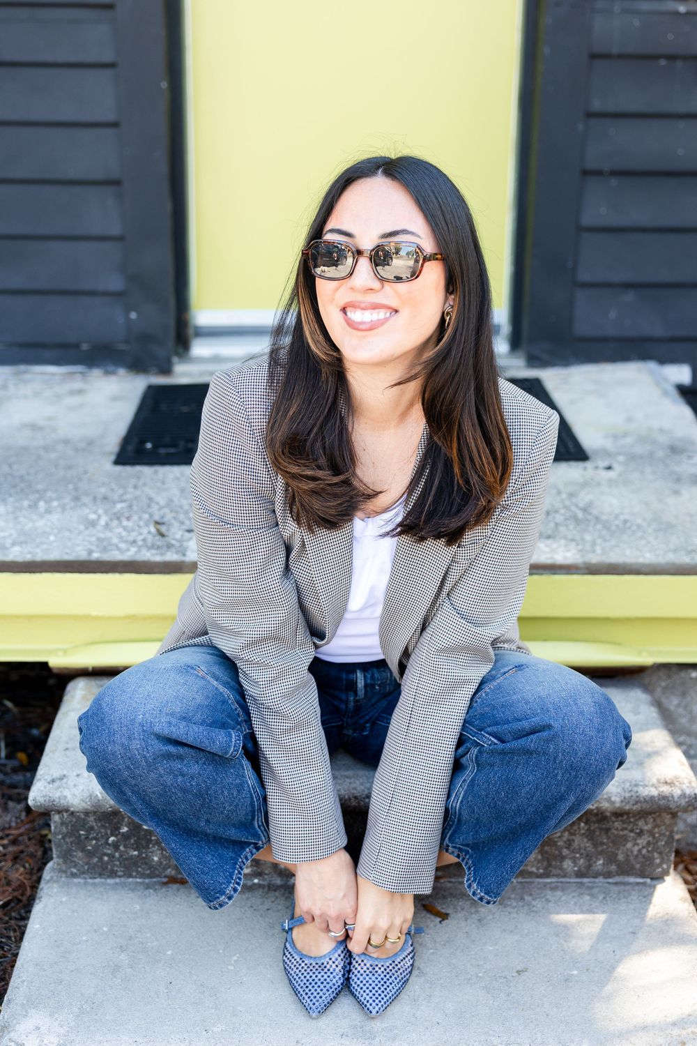 Smiling woman in gray blazer, jeans, and sunglasses sitting on steps in front of a yellow door.