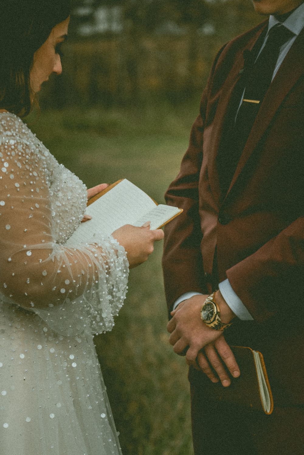 Close-up of hands and open book at outdoor wedding ceremony with bride in beaded dress and groom in dark suit.