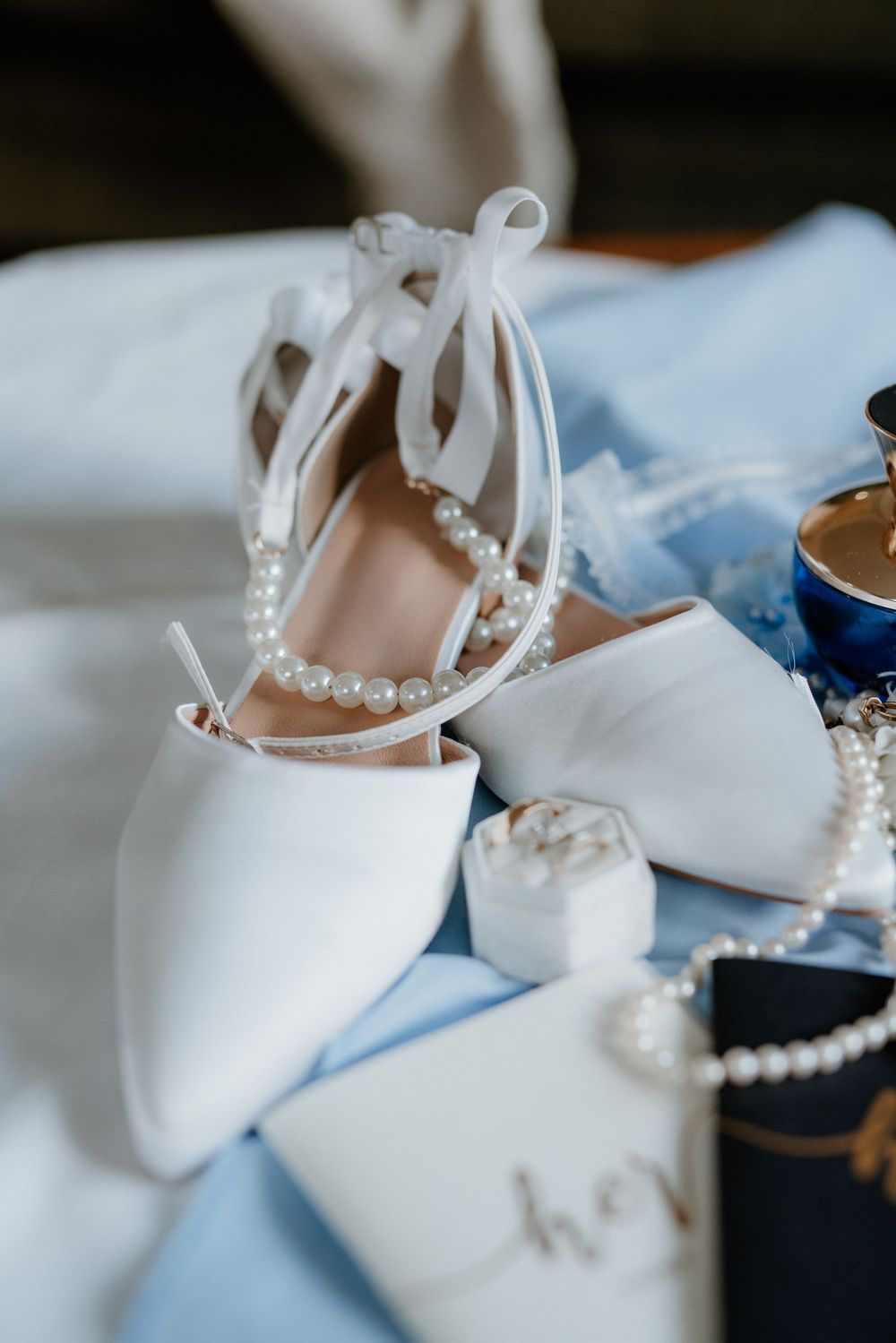 White bridal shoes with pearl straps and wedding accessories arranged on light blue fabric.
