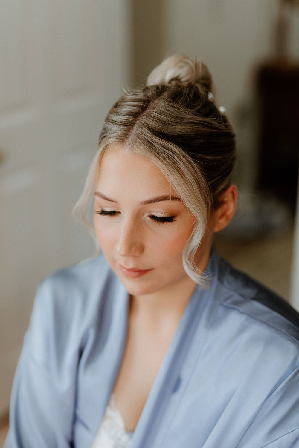 A bride getting ready in a light blue robe with elegant makeup and an updo hairstyle.