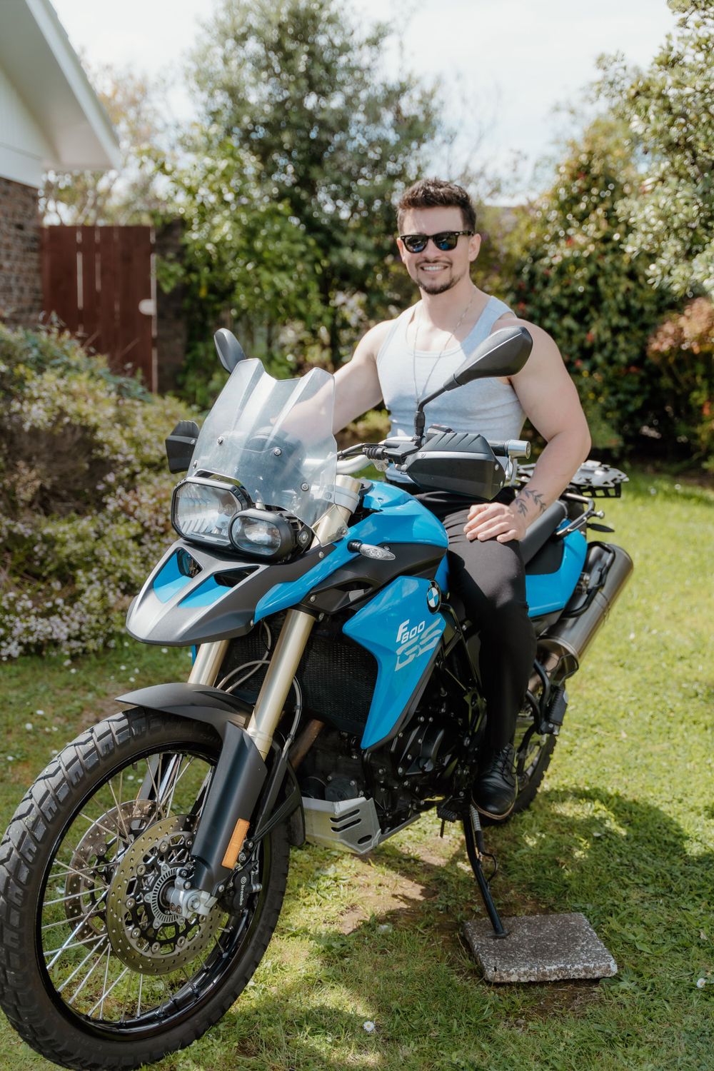 Adventure motorcyclist poses with blue BMW touring bike in garden setting.