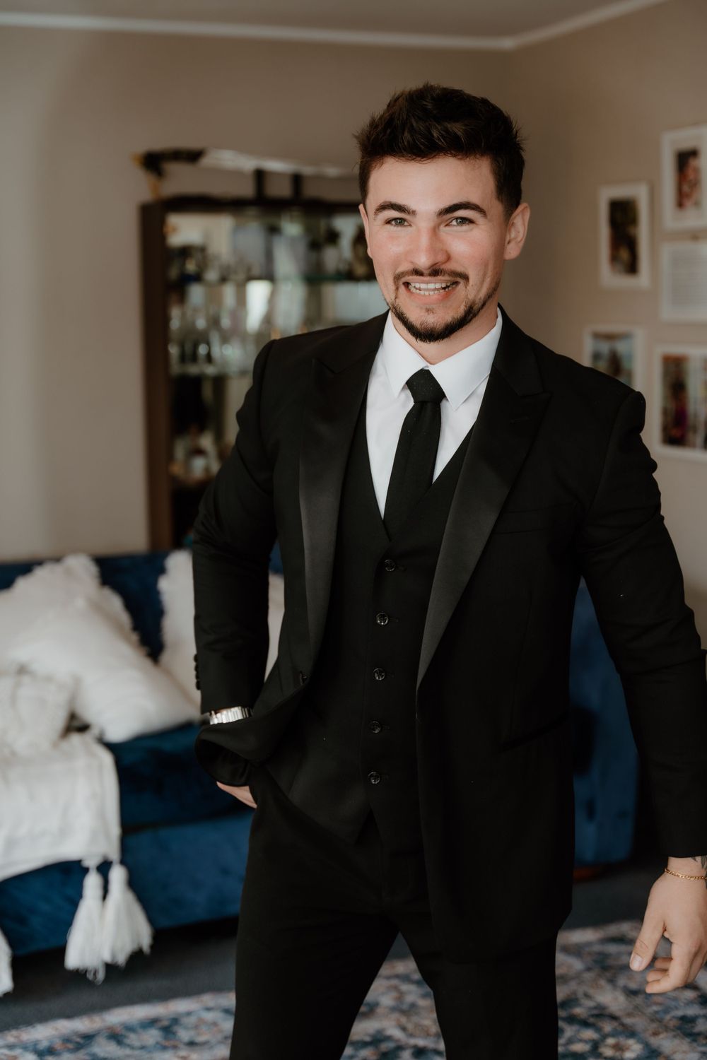 Well-dressed man in black three piece suit and tie smiling in formal indoor setting.