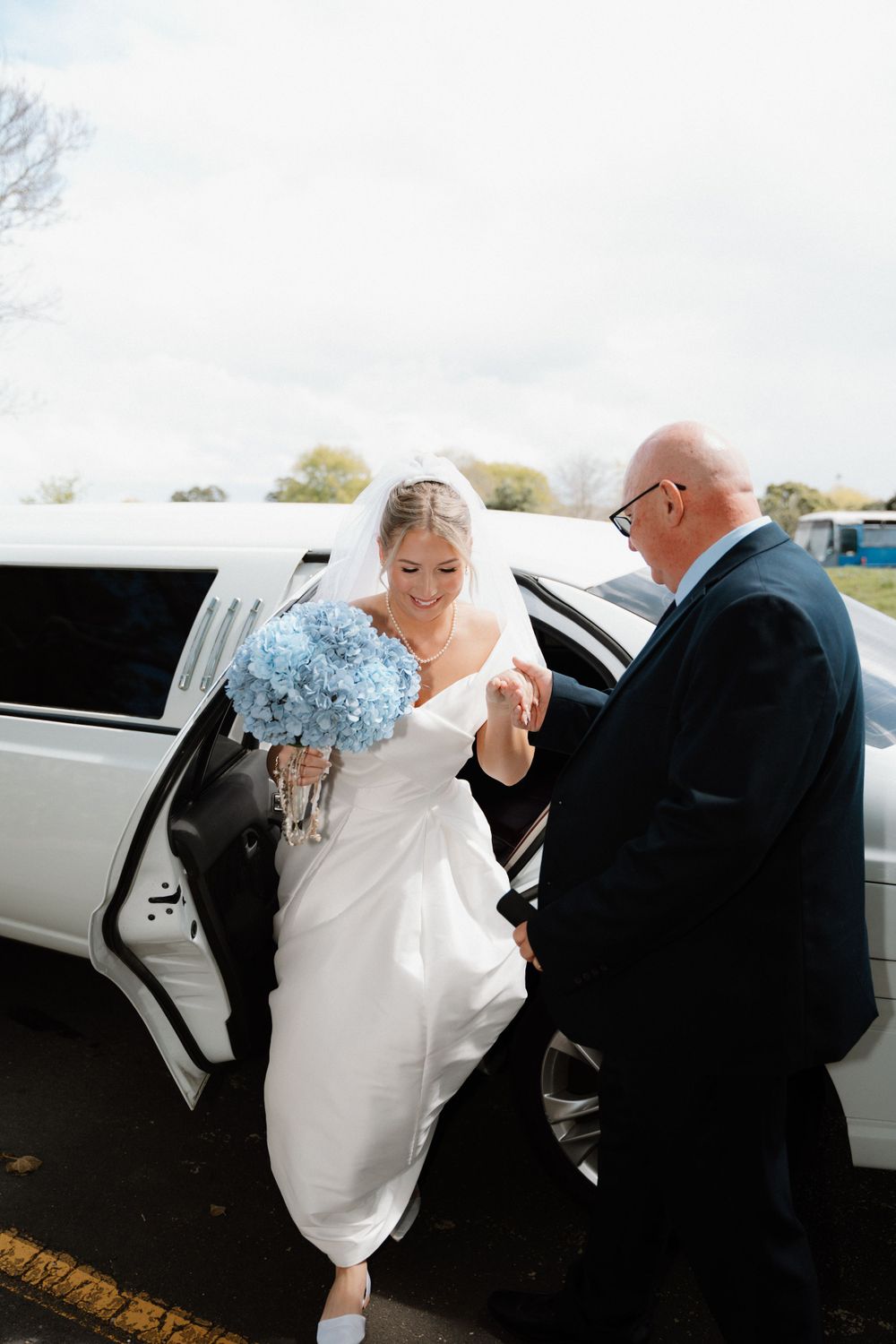 A bride carrying blue flowers exits a limousine assisted by a man in formal attire.