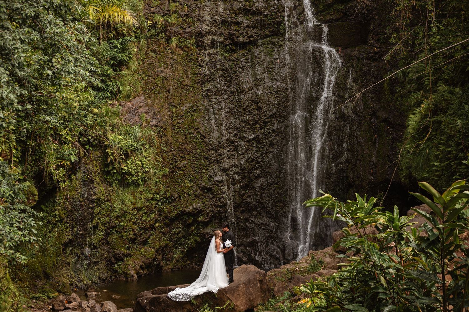 Bride and groom embracing romantically in front of a stunning tropical waterfall surrounded by lush green foliage.