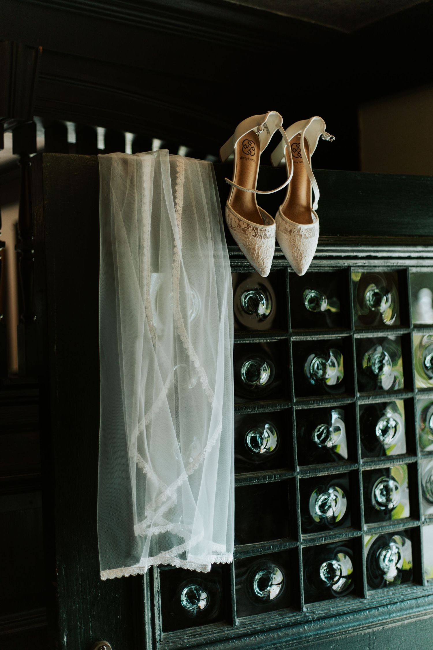 Bridal veil and lace heels displayed on a dark vintage wine cabinet in moody wedding preparation setting.